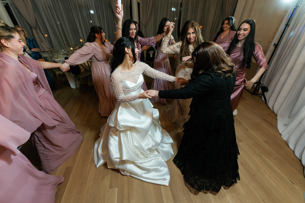 At a wedding reception, a woman in a white gown dances with guests in pink dresses as they hold hands and turn in a circle under warm event lighting.
