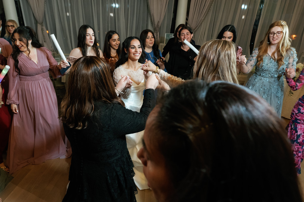 Bride smiling in the middle of a group dancing at a celebration, surrounded by guests in formal attire in an indoor venue.