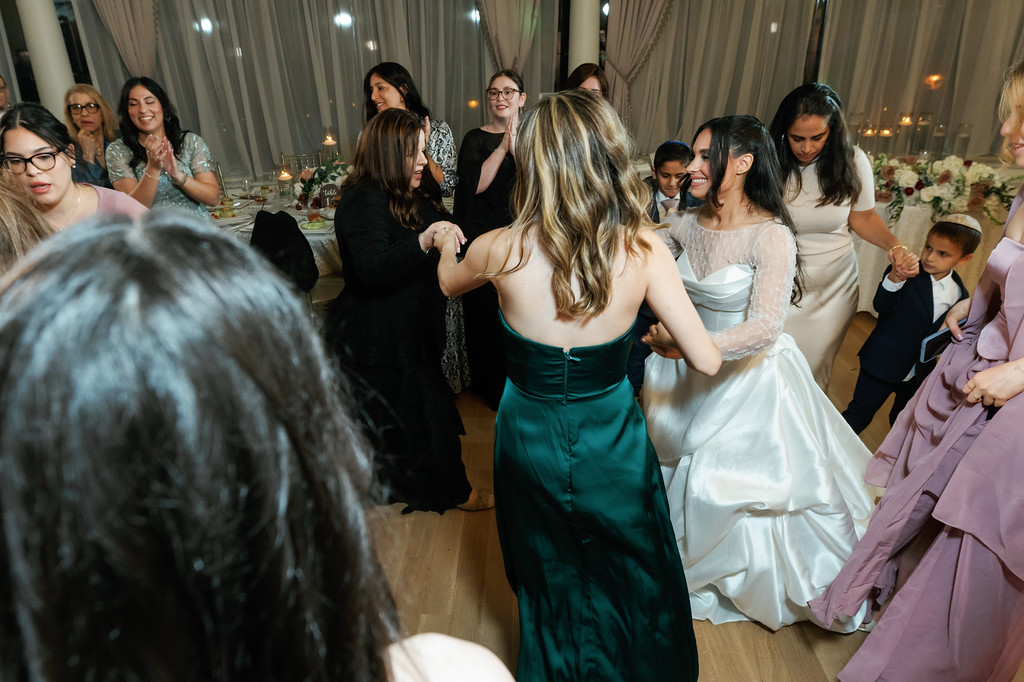 The bride dances with guests at a wedding reception under warm chandelier lighting, with other guests smiling and watching from nearby tables.