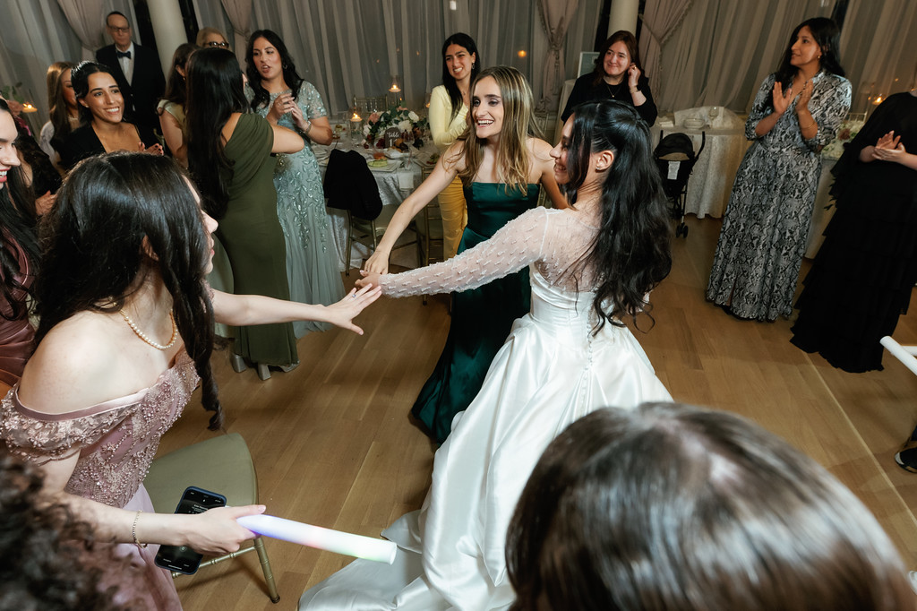 Wedding guests dance in a banquet hall as the bride smiles while others extend their hands toward her in a joyful moment.
