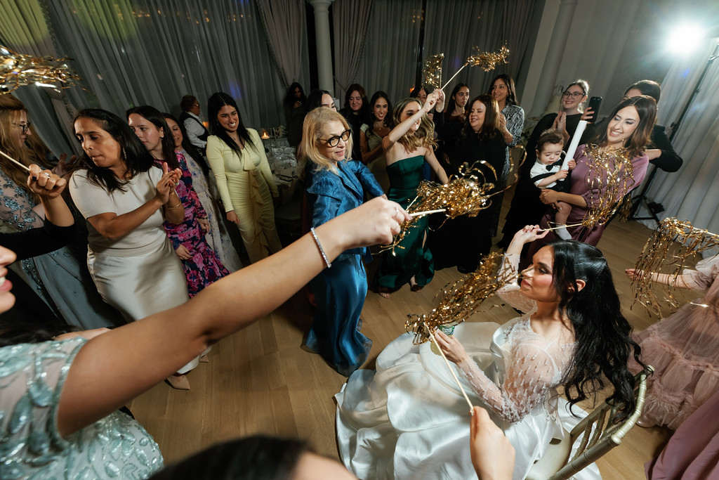 Guests dancing at a wedding reception as the bride sits in the foreground holding a sparkler wand while others smile and raise similar props in a decorated hall.