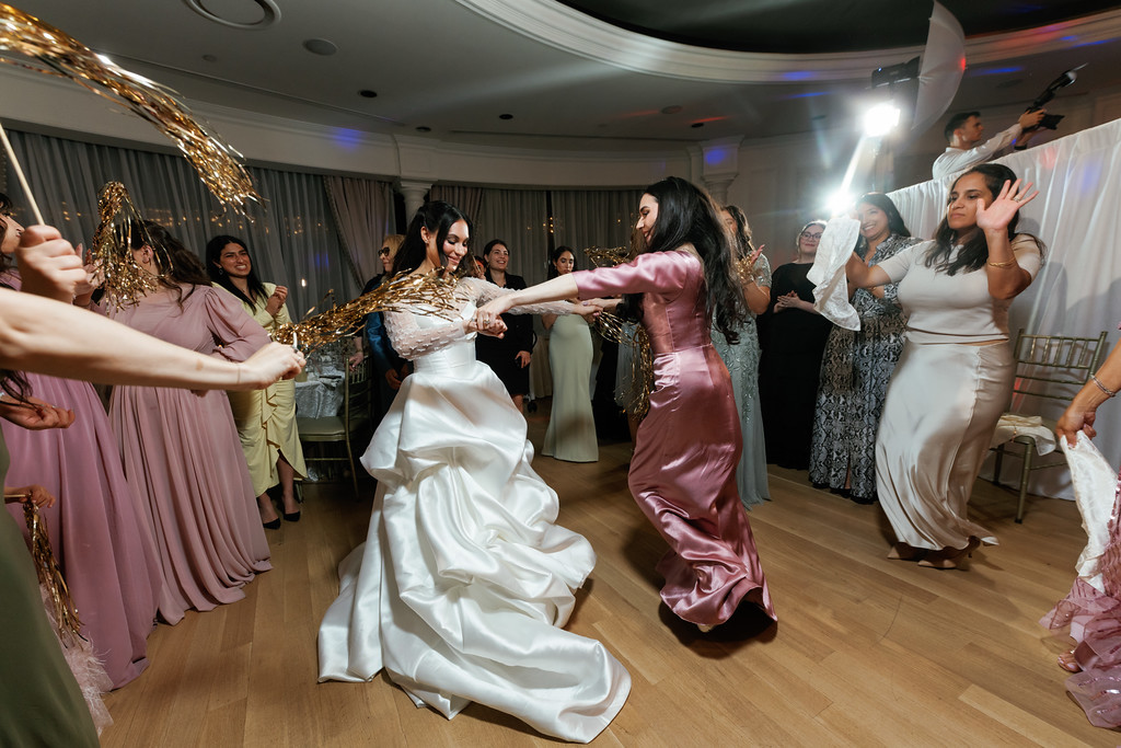 The bride dances on the reception floor with guests, surrounded by gold tinsel decorations and event lighting.