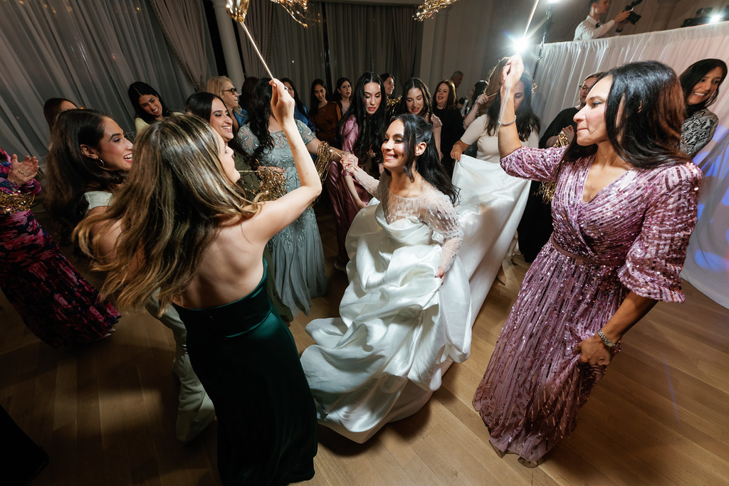Bride in a white dress dancing joyfully with guests at a wedding reception, smiling as people clap and celebrate on the dance floor.