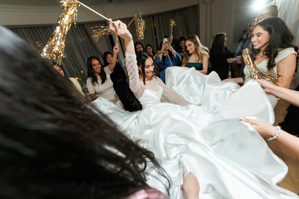 Bride in a white gown smiling while dancing at the reception as guests raise sparklers and streamers in a bright indoor celebration.