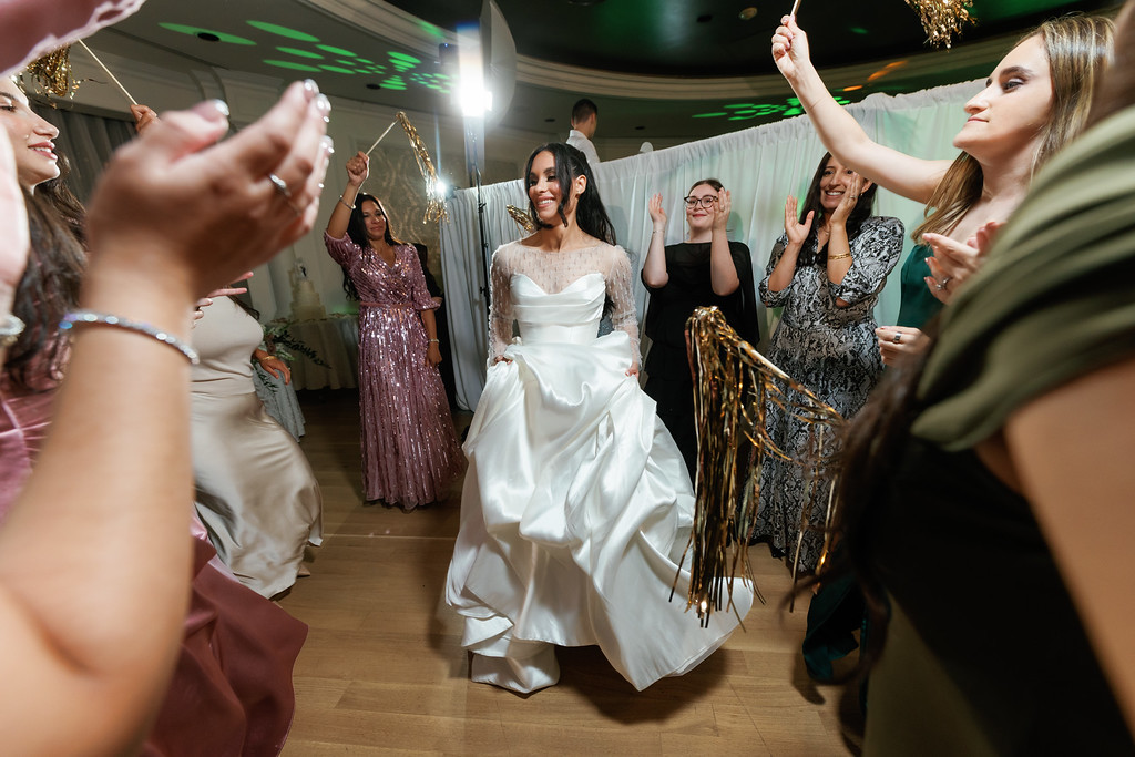 Bride in a white wedding dress smiling and walking through a crowded dance floor as guests clap and cheer, holding strands of gold tinsel at a reception.