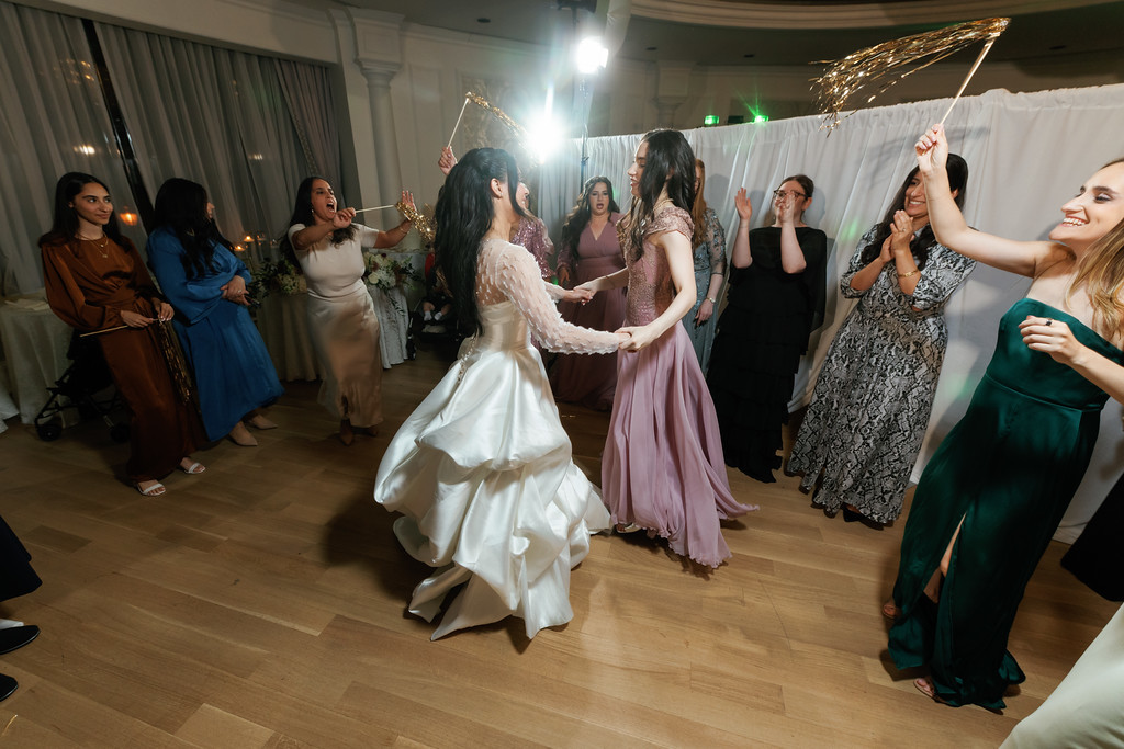 Bride in a flowing white dress dancing with guests at a wedding reception, with bright stage lights and the groom beside her, smiling as everyone celebrates.
