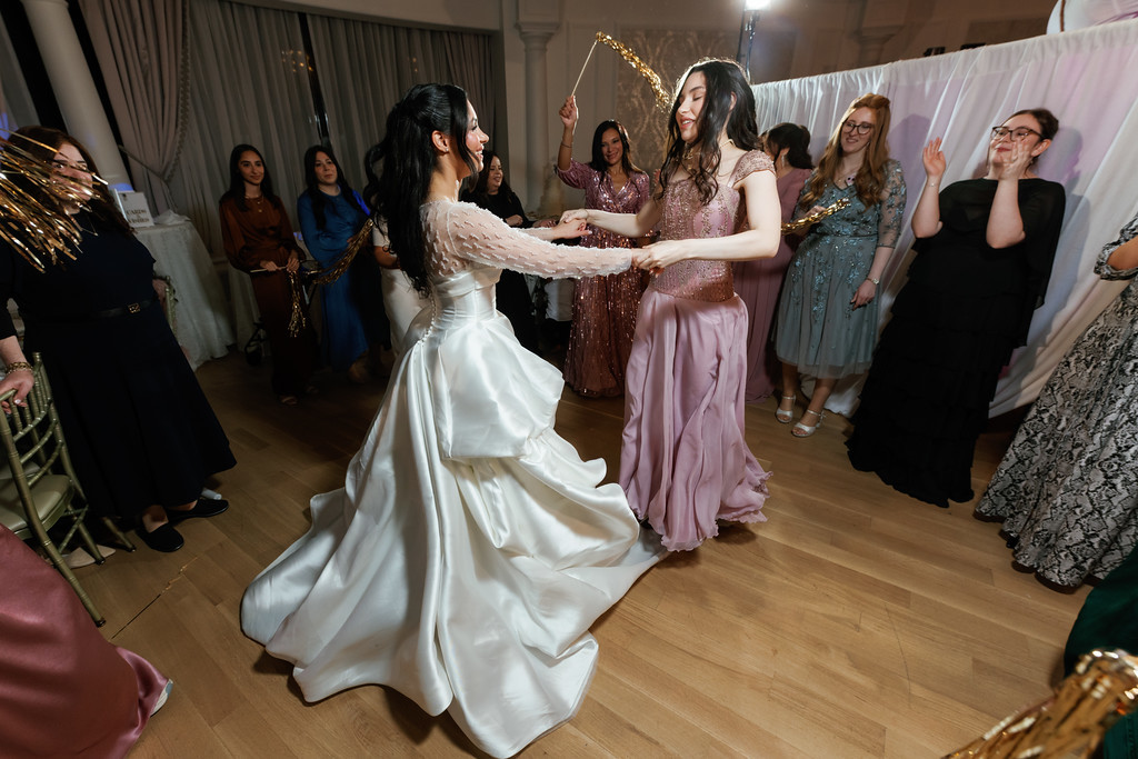 Wedding dance floor moment: a bride in a white dress spins and extends her hands toward another woman in a pink dress, with guests clapping and watching in the background.