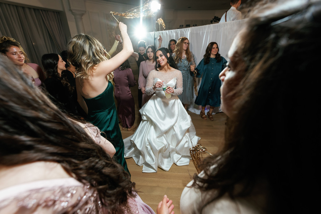 Bride dancing at a wedding celebration indoors, smiling as friends surround her and guests raise their arms in the background.