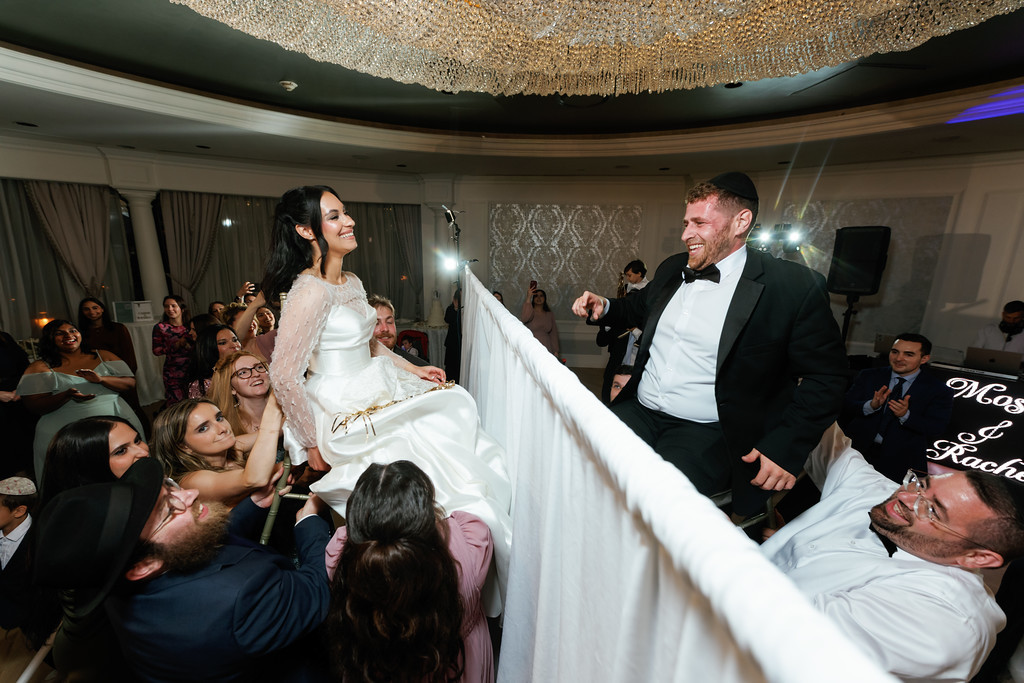 Smiling bride in a white gown and groom in a black tux share a joyful dance moment amid guests at a Jewish wedding reception, with the couple framed by raised arms and a festive backdrop.