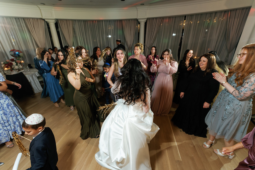 Frum wedding couple dancing with guests surrounding them at a reception, with the bride in a white gown smiling as people clap and celebrate.
