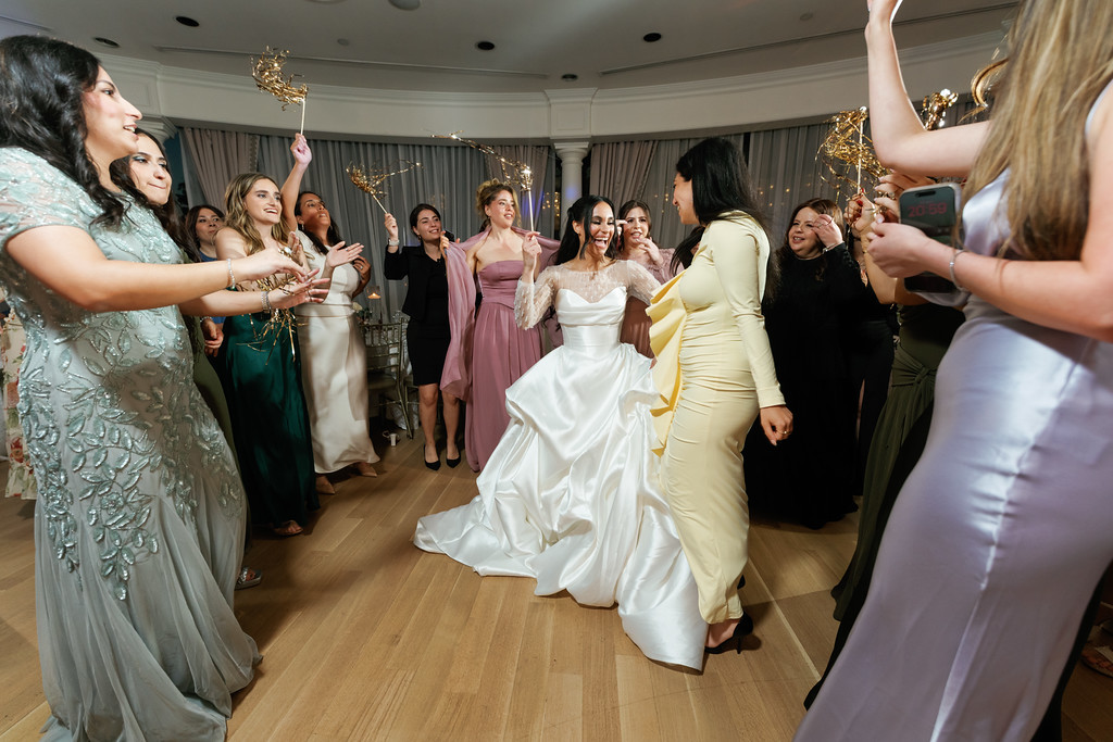 Bride in a white wedding dress twirls while guests dance and cheer at a reception hall, with several women smiling and raising their hands around the dance floor.