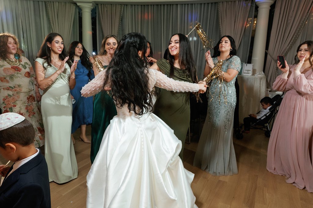 Bride in a white gown dances with her back to the camera while surrounded by women cheering and holding gold tinsel and sparklers at a wedding celebration.