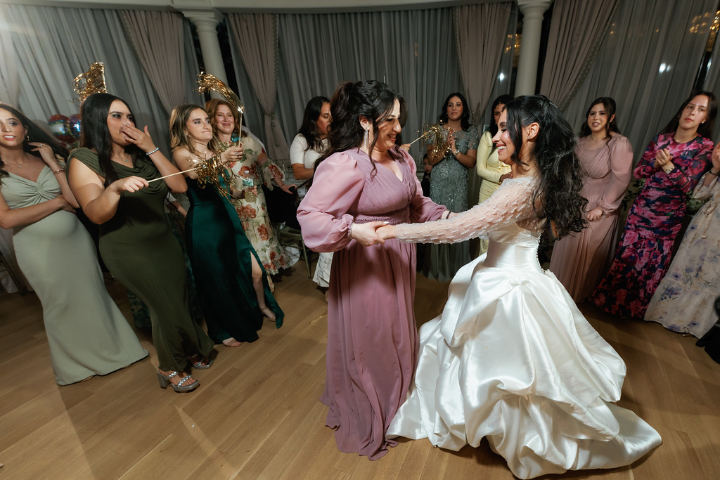 The bride in a white gown dances and smiles with guests at a wedding celebration, with other guests standing along the sides in evening light.