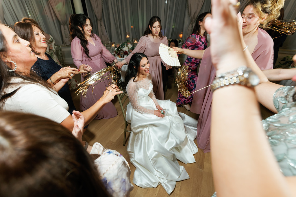The bride smiling while dancing at a wedding reception, surrounded by guests clapping and holding small ceremonial fans under event lights.
