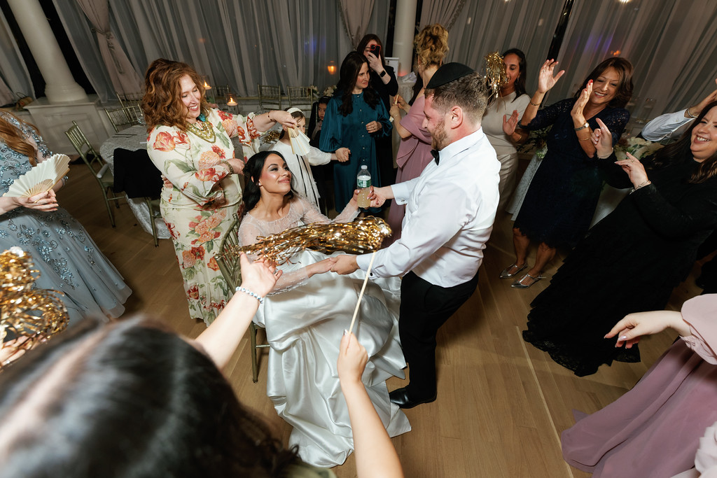 Bride and groom dancing with guests at a Jewish wedding reception under tented lights, the bride smiling in the foreground as nearby guests celebrate with raised hands.