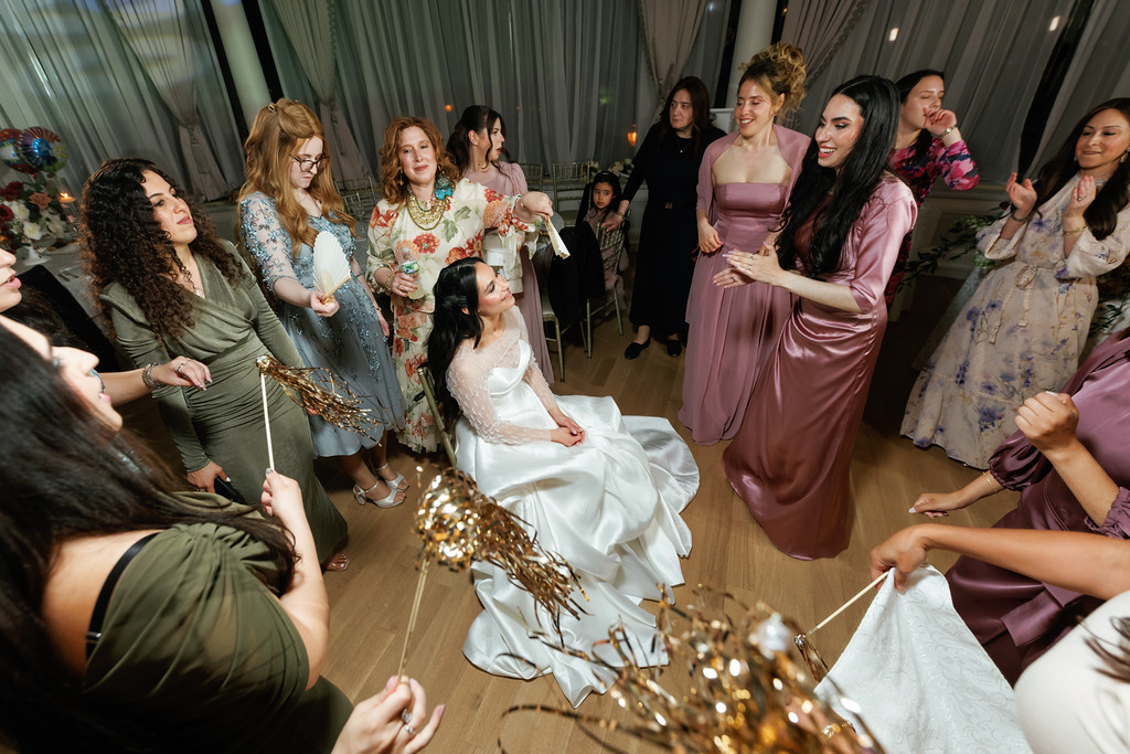 The bride dances with friends as women surrounding the party clap and smile during a wedding celebration under a draped canopy.
