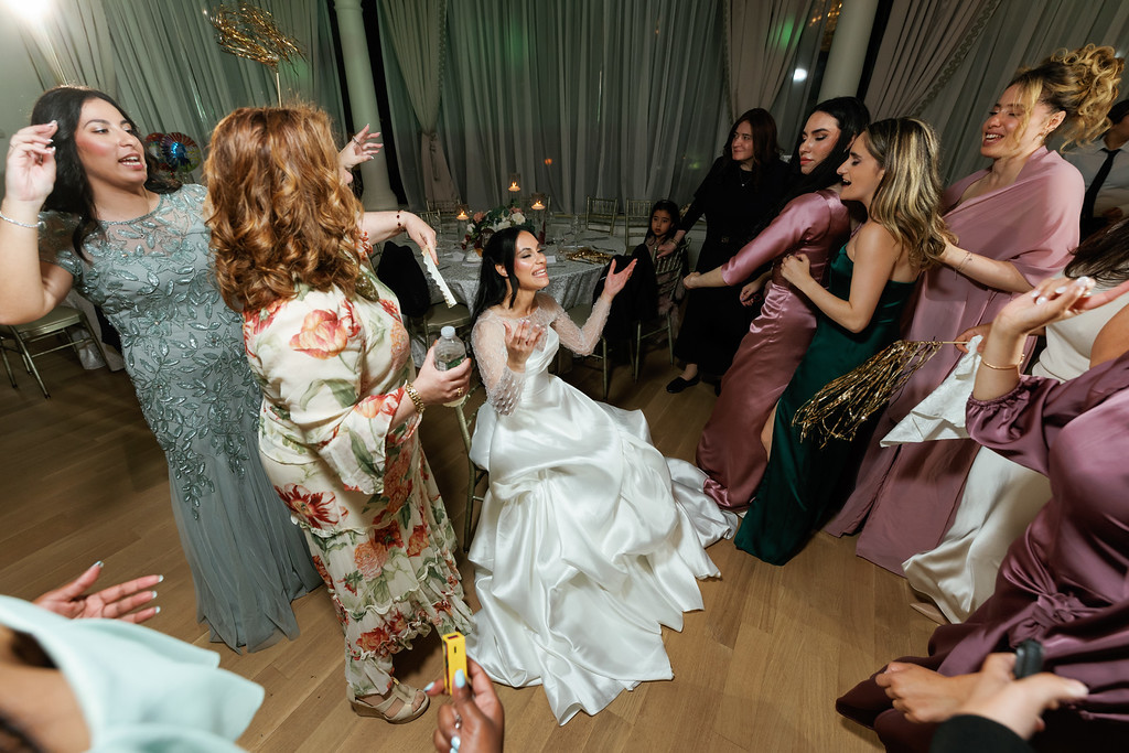 A group of women and guests dance and clap at a wedding reception while the bride in a white gown moves through the crowd, smiling and celebrating.