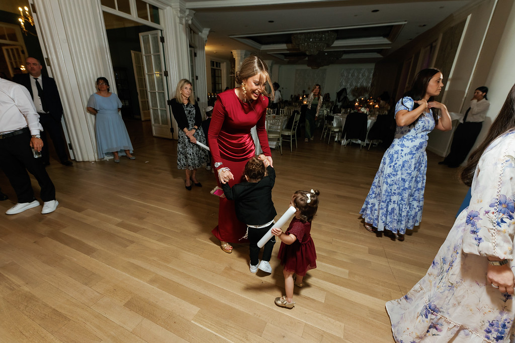 Bride in a red gown dancing with a young child on a wedding reception floor, with other guests in formalwear in the background.