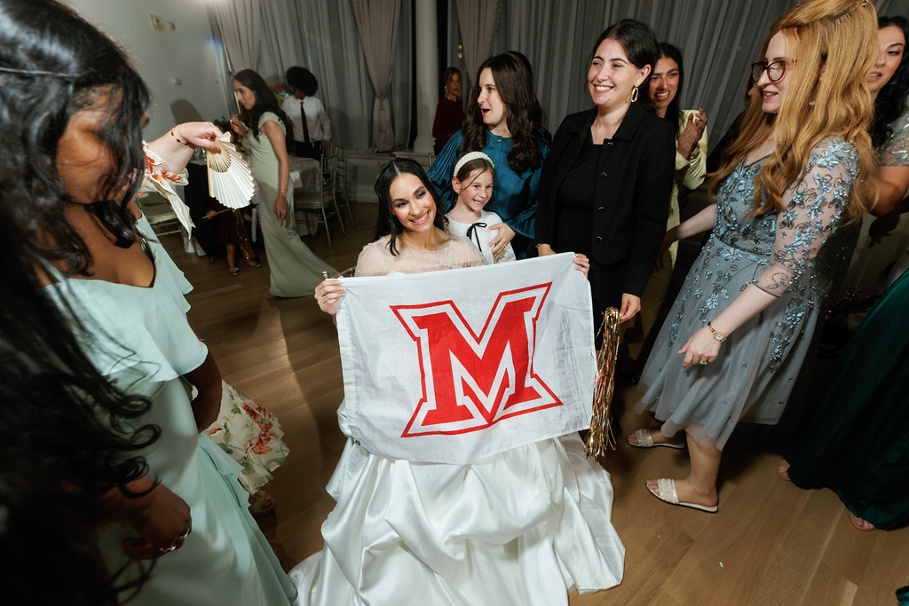 Bride holding an “M” banner while smiling during wedding party dancing, with friends surrounding her in a banquet hall.