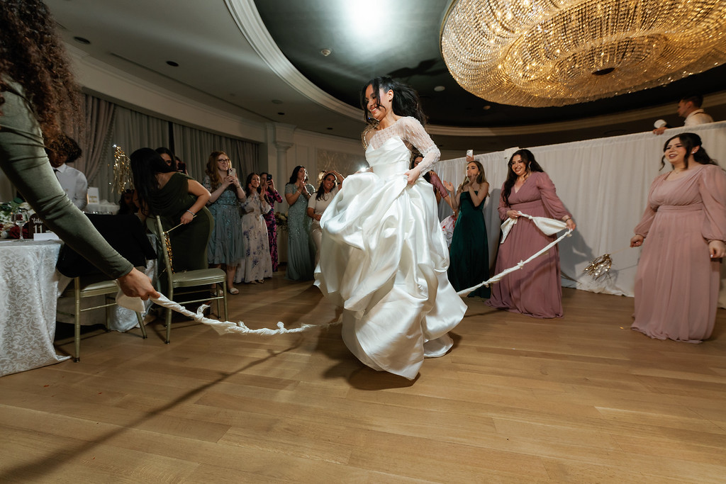A bride in a white gown spins while dancing at a wedding, with guests clapping in the background under warm ballroom lighting.