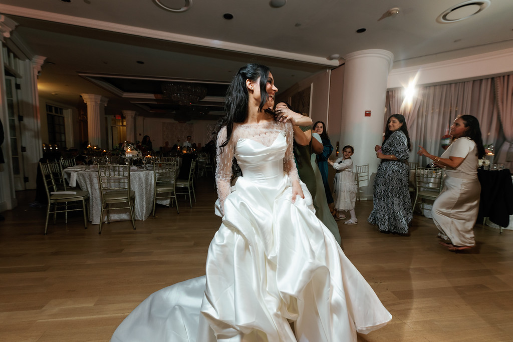 Frum bride in a white wedding gown dancing and looking toward a cheering moment as guests dance in the background inside the hall.