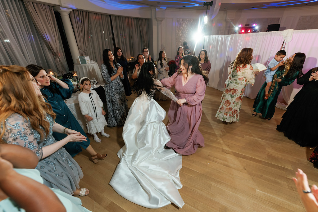 At a Jewish event, the bride dances on the floor while guests in formal attire surround her, smiling and watching near a stage area.