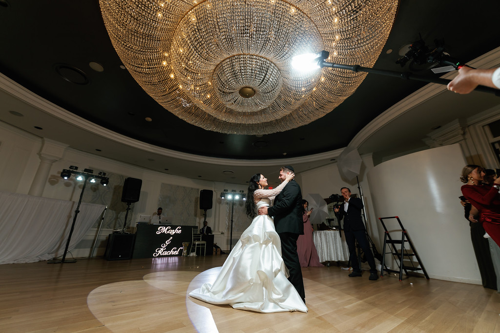 A bride and groom embrace while dancing in a ballroom decorated with a large sparkling chandelier overhead.