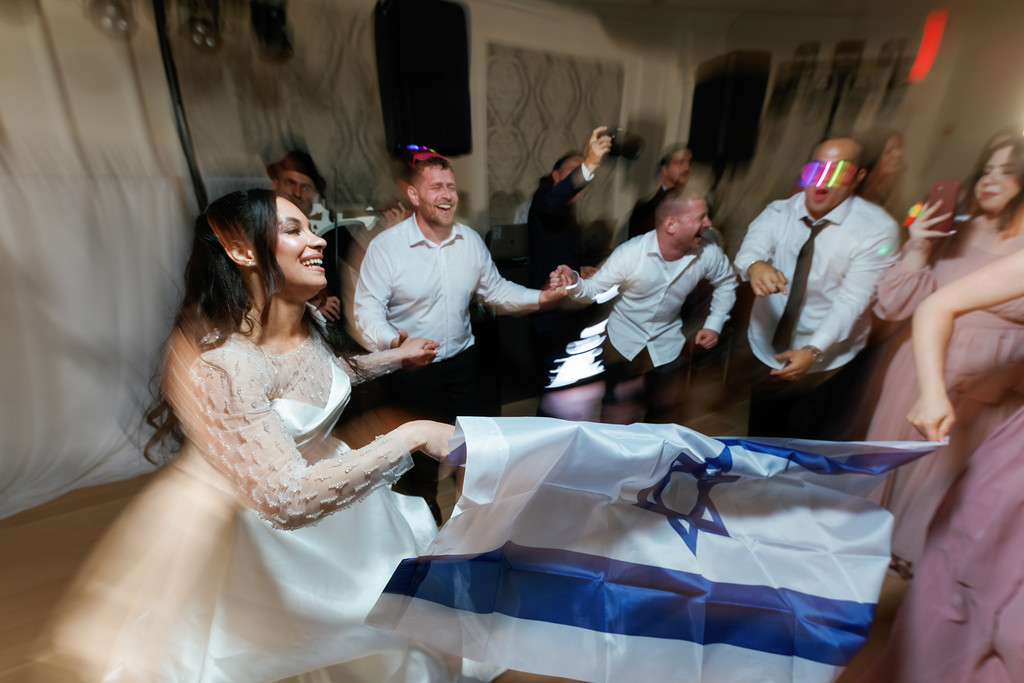 A joyous bride in a white gown dances in front of a crowd holding a blue-and-white Torah flag, smiling as guests celebrate behind her.