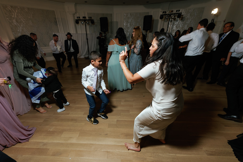 Guests dance at a wedding reception while a young boy in a white shirt and vest moves through the center of the dance floor.