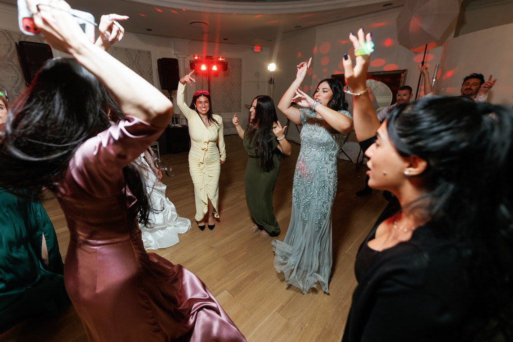 A joyful group dances at a wedding reception as the bride in a light blue dress raises her hands, with guests cheering and moving around a dance floor under red and green lights.