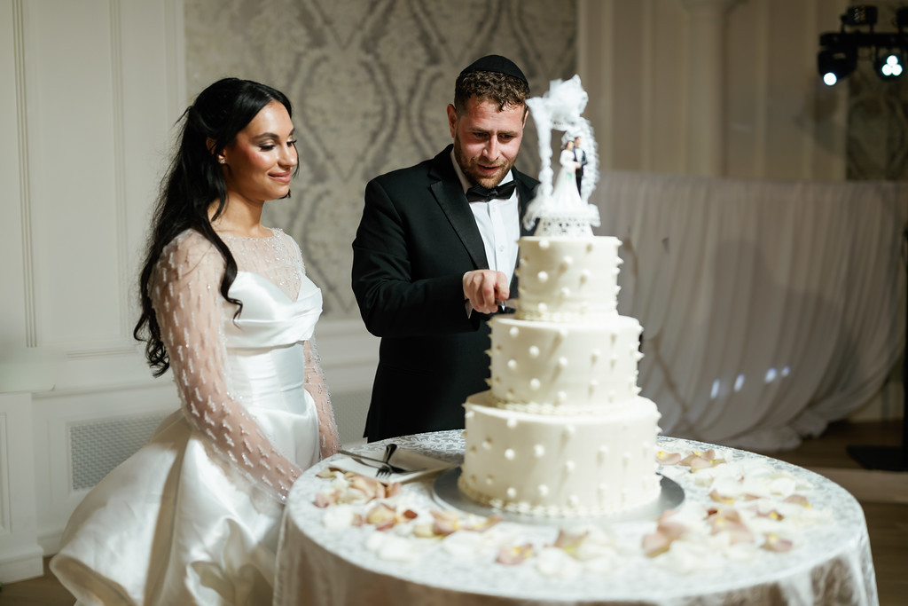 Bride and groom in formalwear cut a multi-tier wedding cake together at a ballroom table, smiling as the groom’s hand reaches the knife.