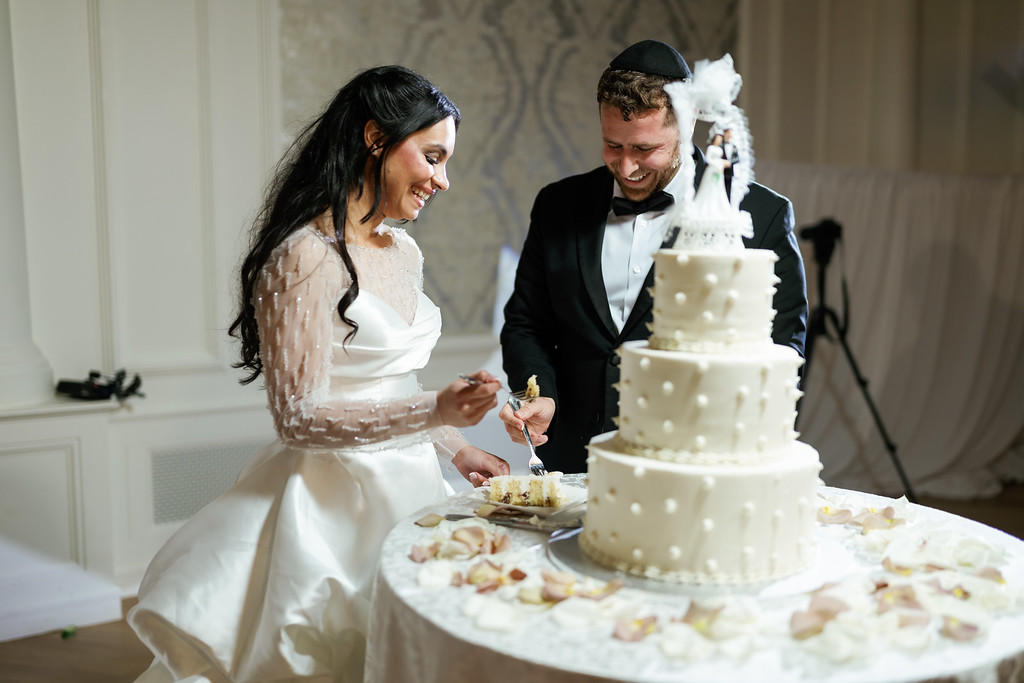 Bride and groom smiling while cutting and tasting wedding cake together at the reception table.
