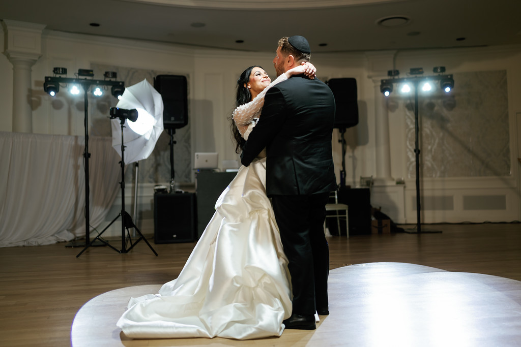 A bride and groom in formal wear embrace and dance in a bright ballroom, the bride smiling with her veil while the groom holds her close.