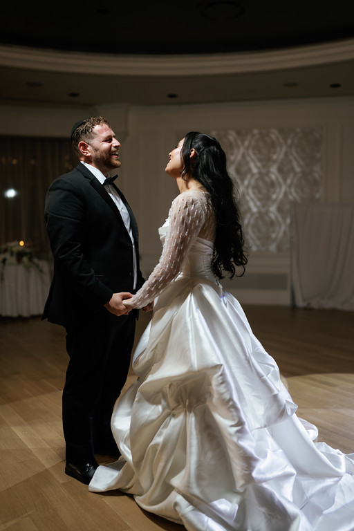 Bride and groom holding hands while facing each other and smiling during a quiet dance in a ballroom.