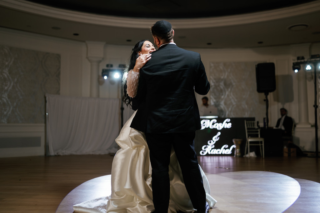 Bride and groom in an intimate dance embrace on the wedding floor, bride’s veil and gown catching the light, with banquet hall stage lights in the background.