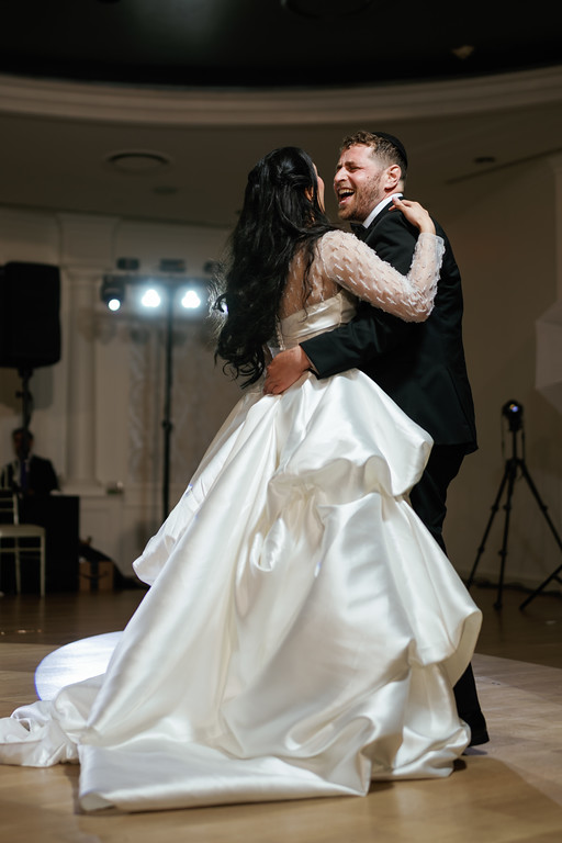 A bride and groom dance together in a ballroom, holding each other and smiling as their embrace turns mid-sway.