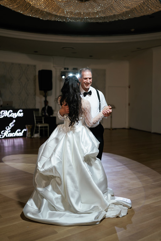 A bride and groom dancing together in a ballroom, the groom smiling while the bride’s white dress flares as they spin.