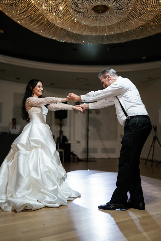 A couple dances at their wedding, extending their hands to each other under a large chandelier in a bright hall.