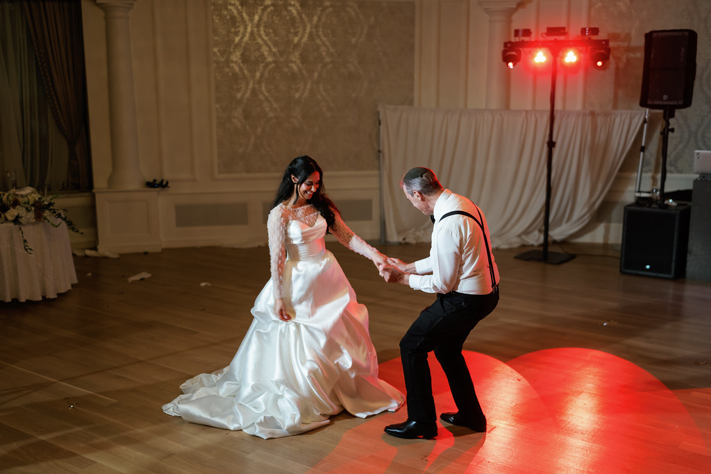 The bride in a white gown and headpiece sits on the dance floor while the groom kneels in front of her holding her hands, with colored stage lighting in the ballroom background.