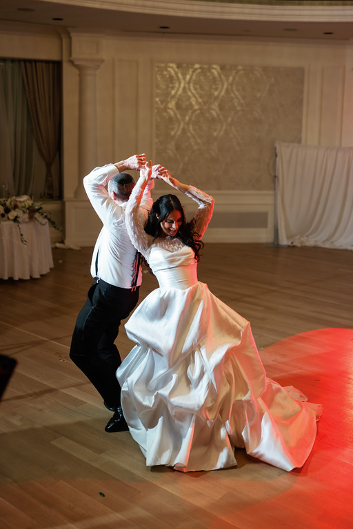 Bride in a white gown and groom in a white shirt and dark pants dance together mid-spin, lifting their arms as the bride’s skirt billows across the wooden ballroom floor.
