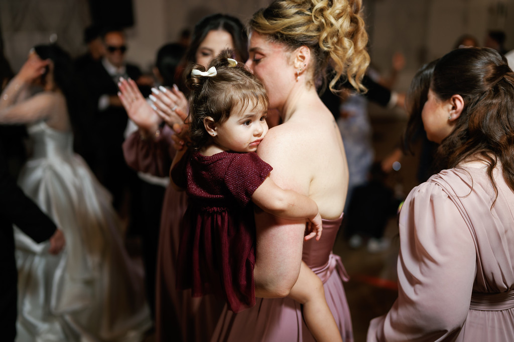 Bride holding her toddler daughter during a joyous dance at a wedding ceremony, with family and guests clapping in the background.
