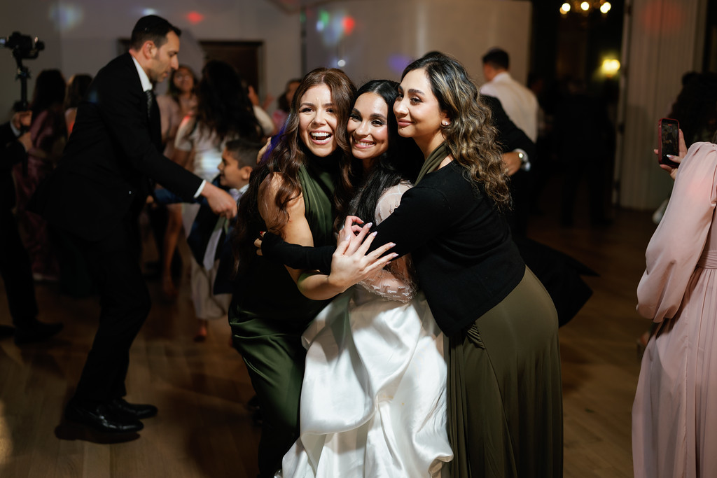 Three women dance together in a close group embrace, smiling brightly at a wedding reception while guests and a smartphone blur in the background.