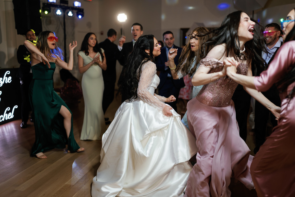 Reception dance-floor moment: guests in formal attire clap and sing as the bride’s group dances, with warm party lighting and colorful highlights in the background.