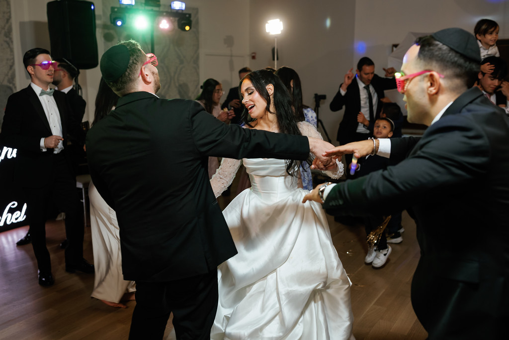 The bride and groom dance closely together on the reception floor, smiling and leaning into a joyful embrace while guests watch in the background.