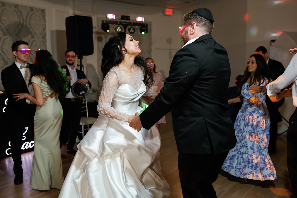 A bride in a white wedding dress and her groom in a black suit dance together at a reception, smiling and turning while guests watch in the background.