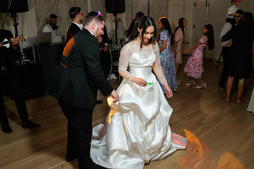 Bride in a white wedding gown and lace sleeves glances down while being guided during reception dancing, with guests dancing in the background under event lighting.
