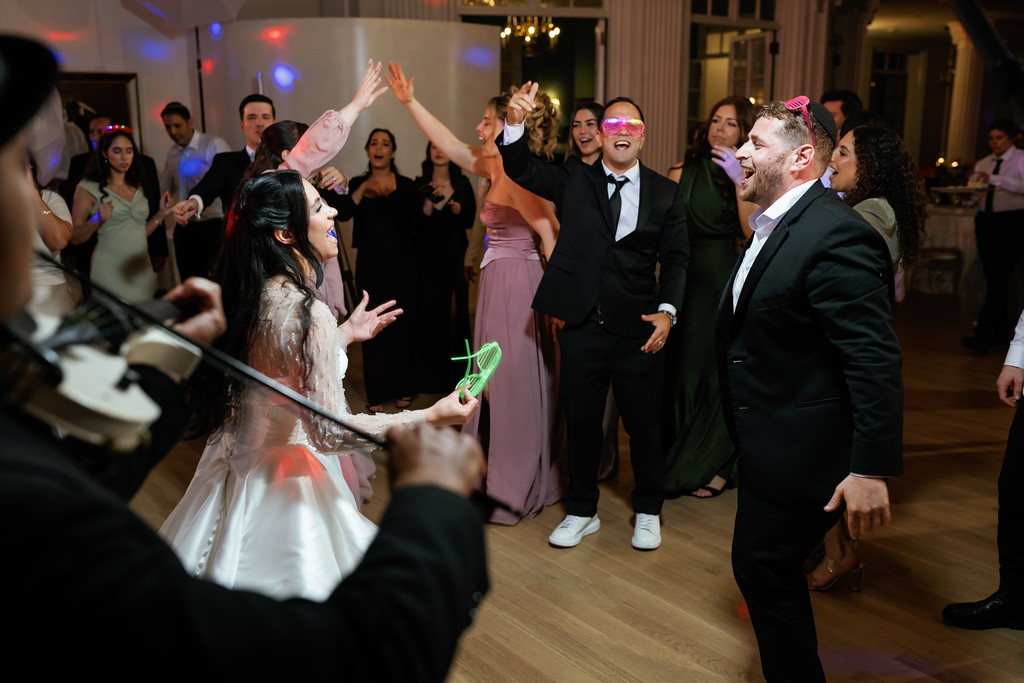 The bride and groom dance at a wedding reception as guests around them clap and reach their hands up in celebration, with colorful event lights in the background.
