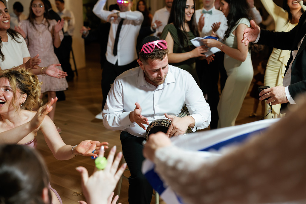 A man wearing a pink hat and kippah dances at a Jewish wedding reception while guests around him clap and smile.