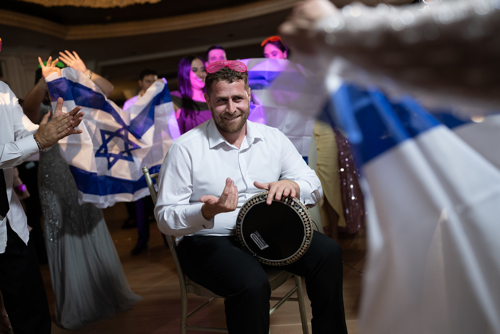 Smiling groom seated while playing a large ceremonial drum, gesturing with his hands as guests in the background dance and wave flags at a wedding reception.