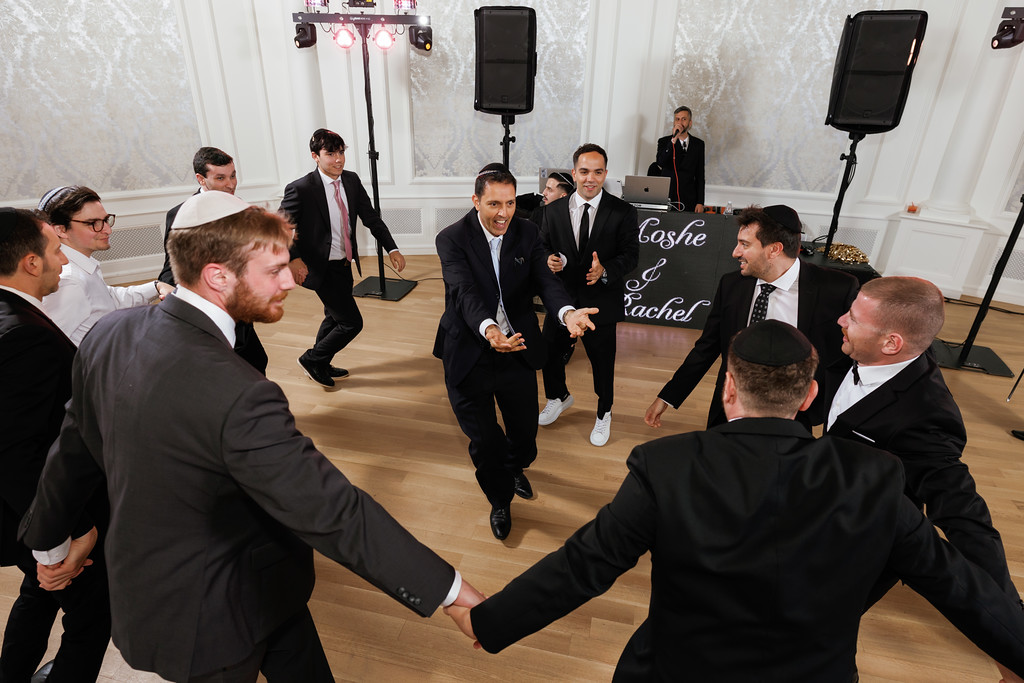 Wedding reception guests dance in a circle, holding hands as the groom and friends smile under event lights.